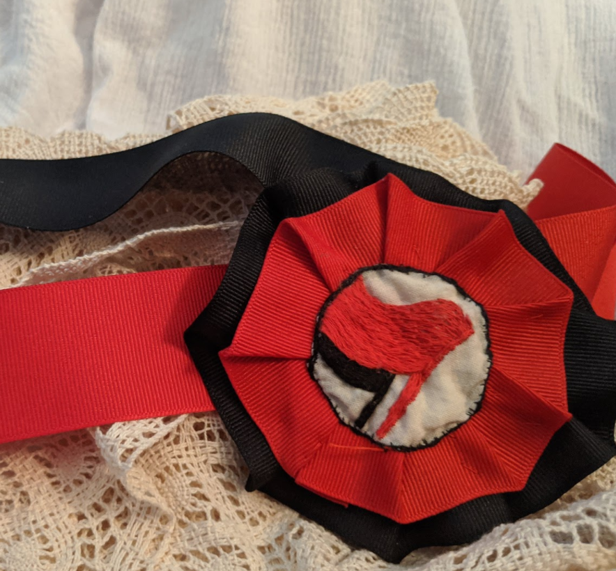 An antifa cockade of black and red ribbon with a red and black antifa flag logo embroidered onto a white piece of fabric. The cockade rests on a piece of white lace.